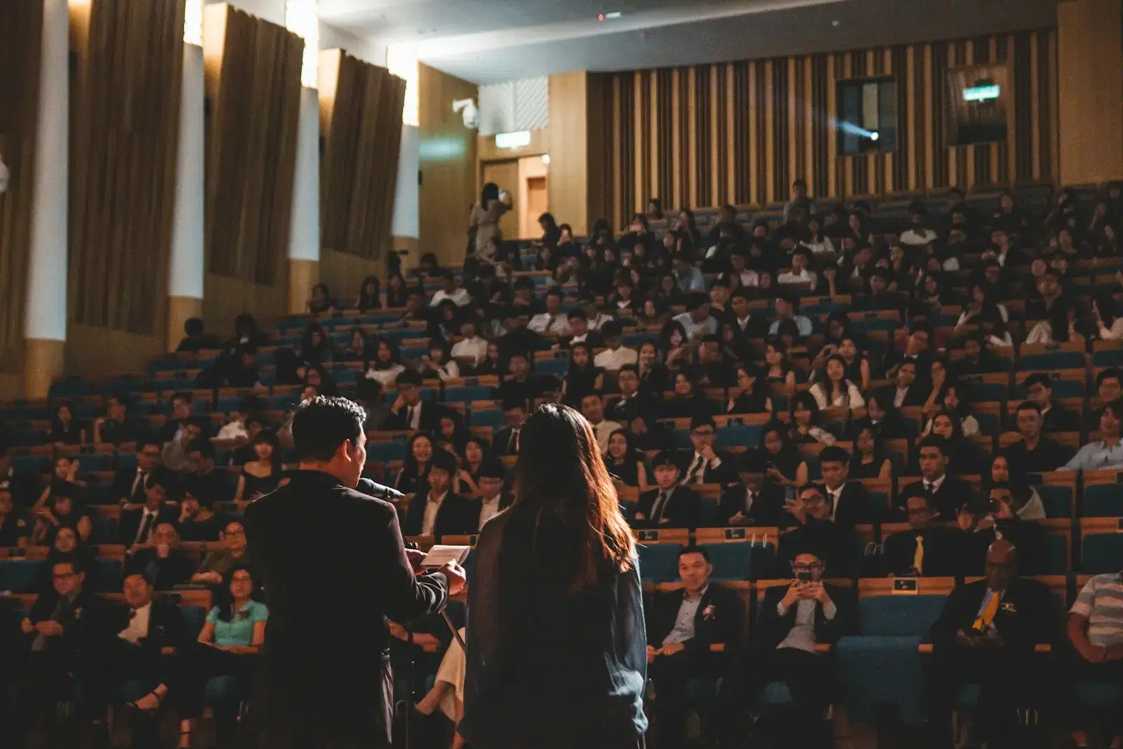 A full auditorium seen from the back, two people standing in the aisle