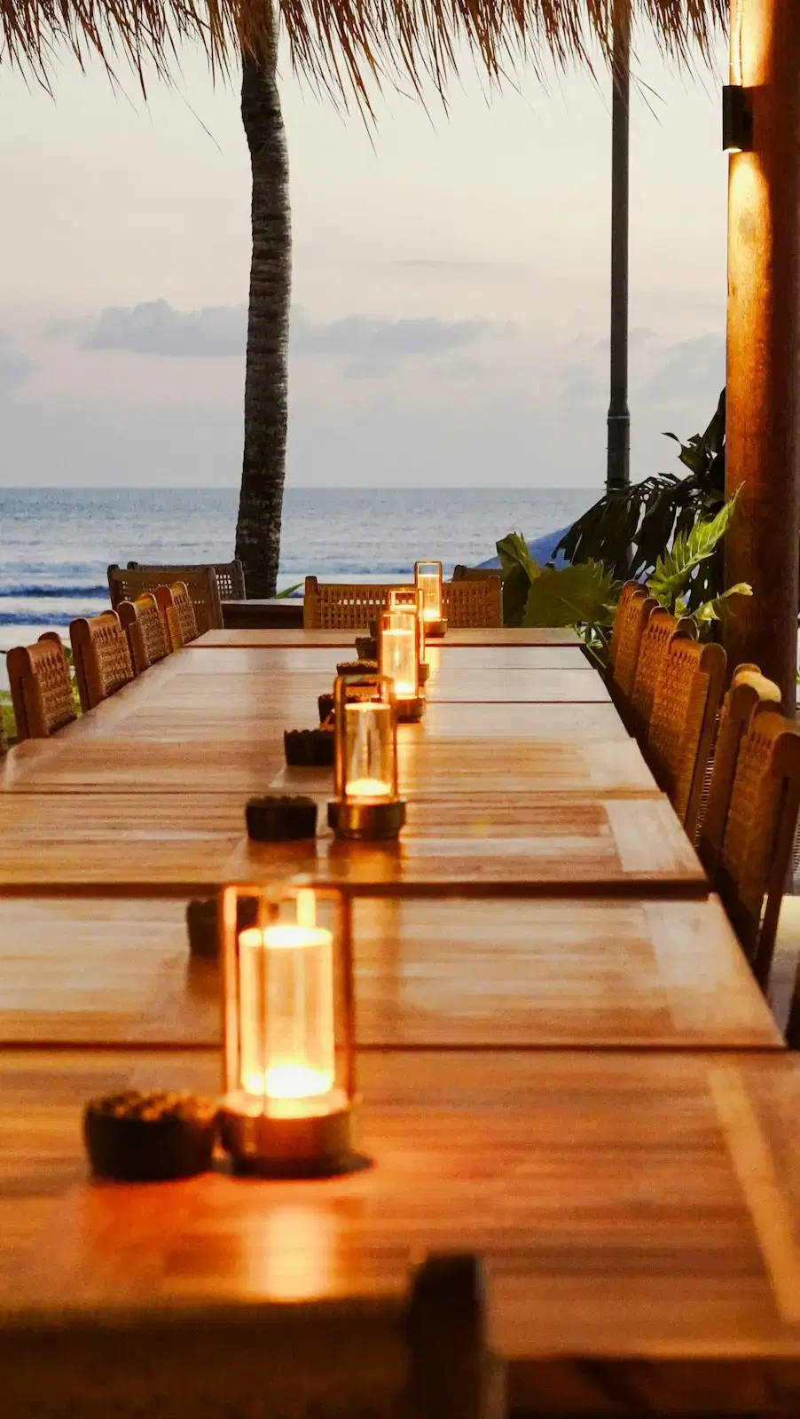 A long wooden table set with lit candle lanterns and rattan chairs, the sea beyond it