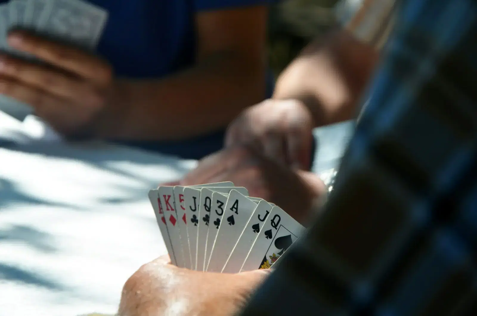 A hand holding a fan of playing cards at a sunny outdoor table, the other players out of focus