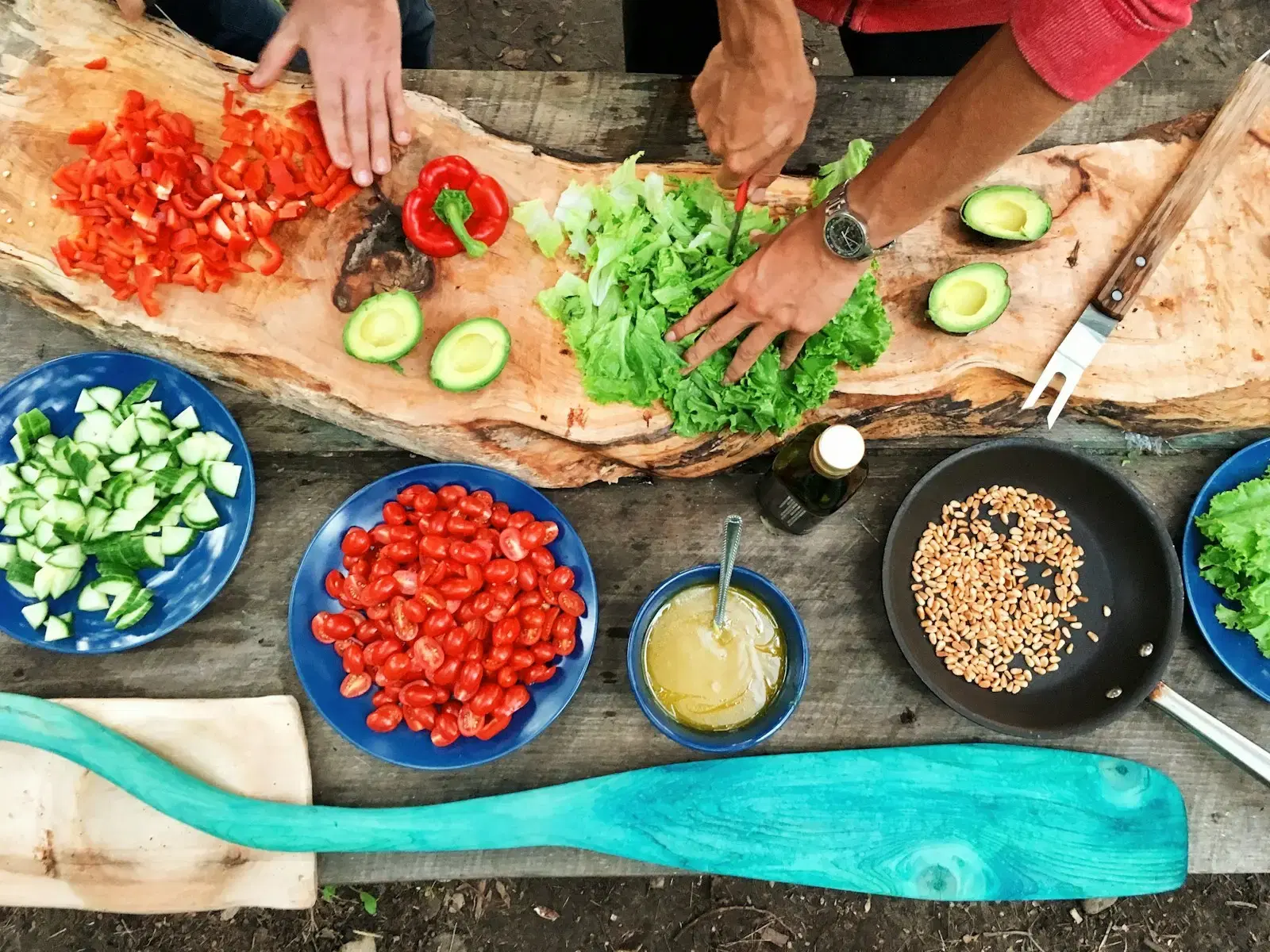 Hands slicing peppers, avocado and lettuce on a wooden board, bowls of chopped vegetables around it