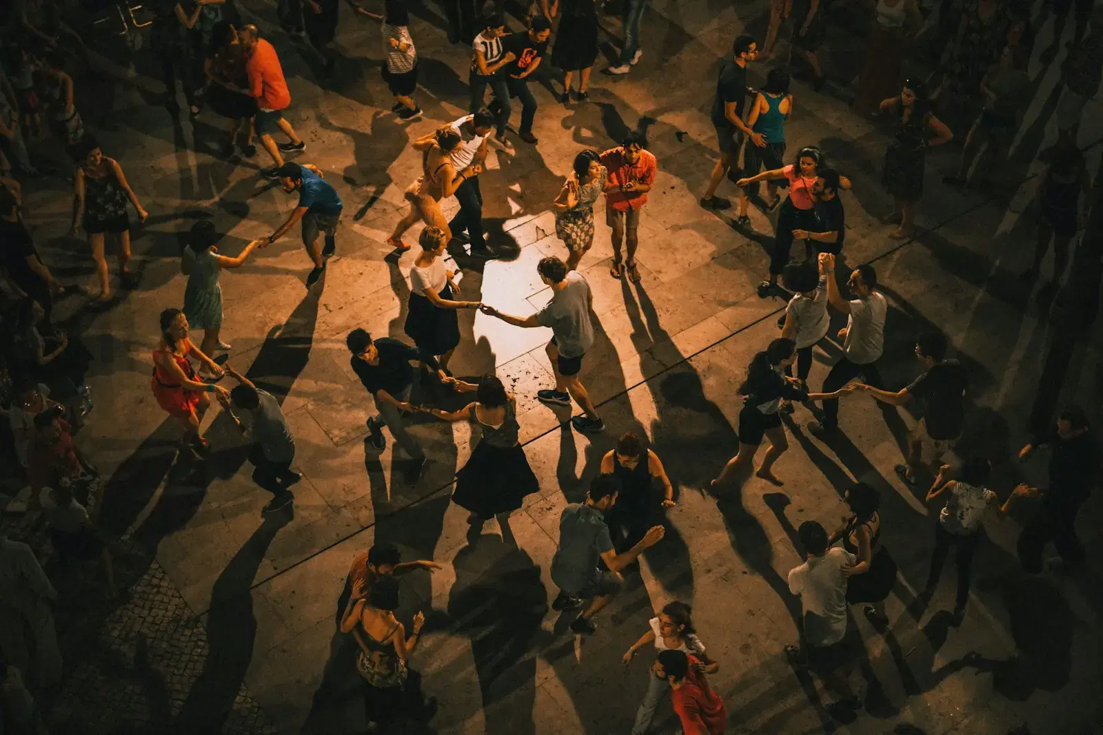 A crowd dancing on a warm-lit floor, seen from a balcony, with long shadows
