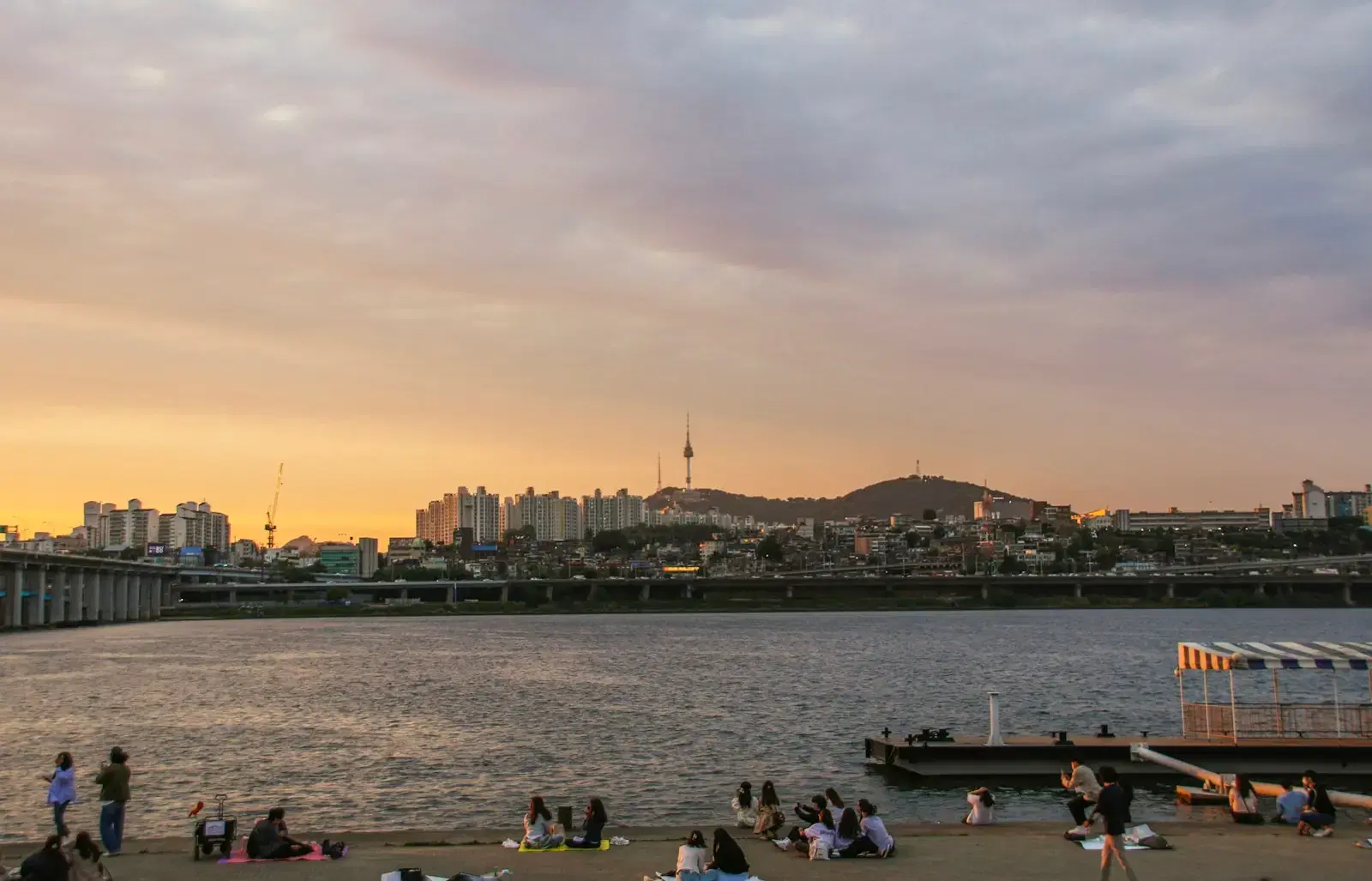 People sitting on a riverbank at sunset, a city and a hilltop tower beyond