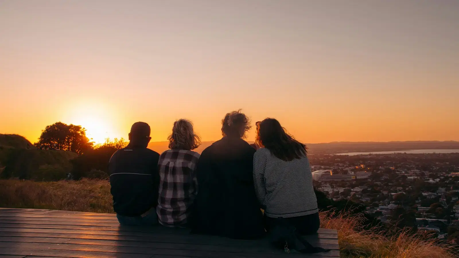 Four friends on a bench on a hilltop, watching the sun set over a city