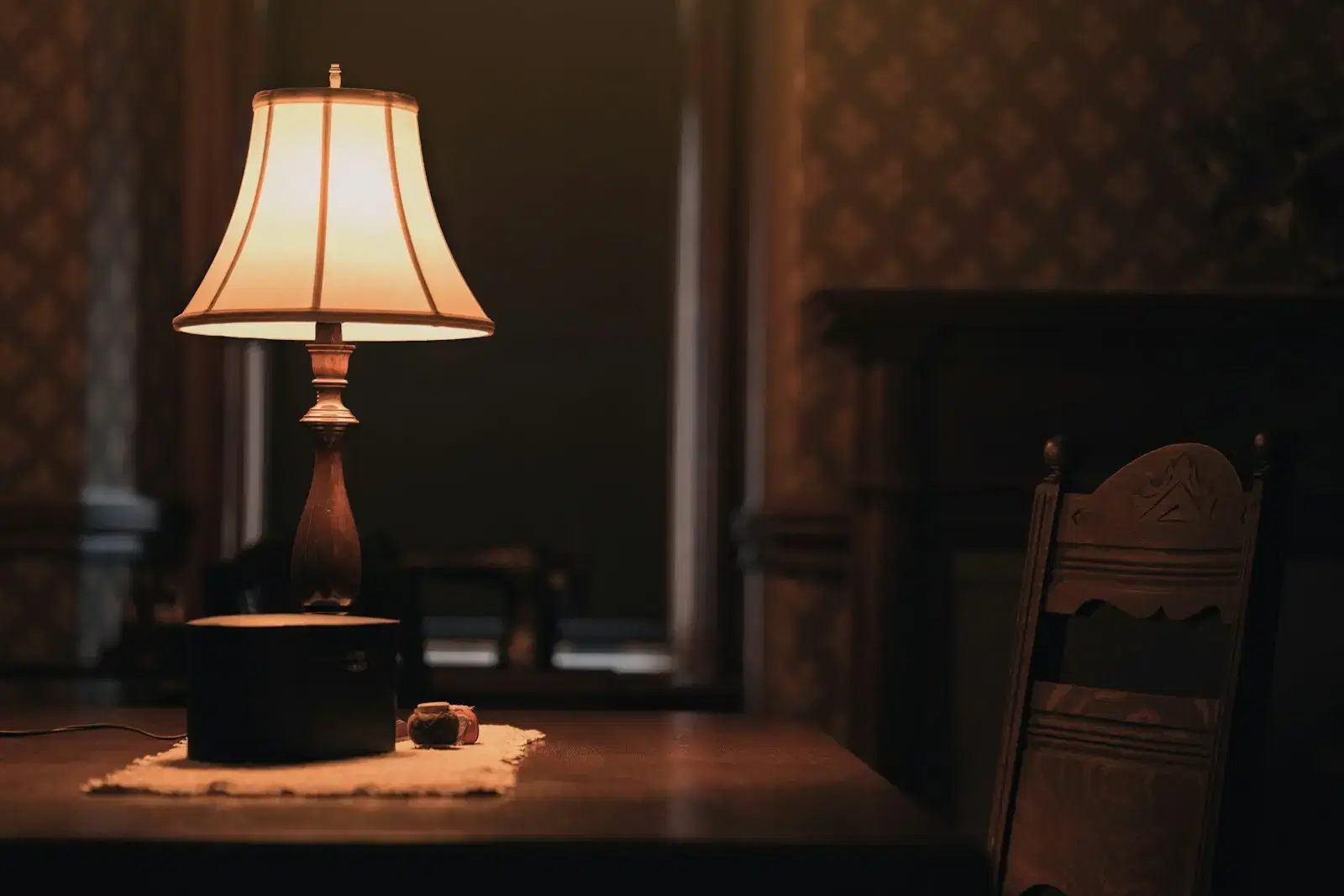 A table lamp glowing on a dark wooden table beside an empty carved chair