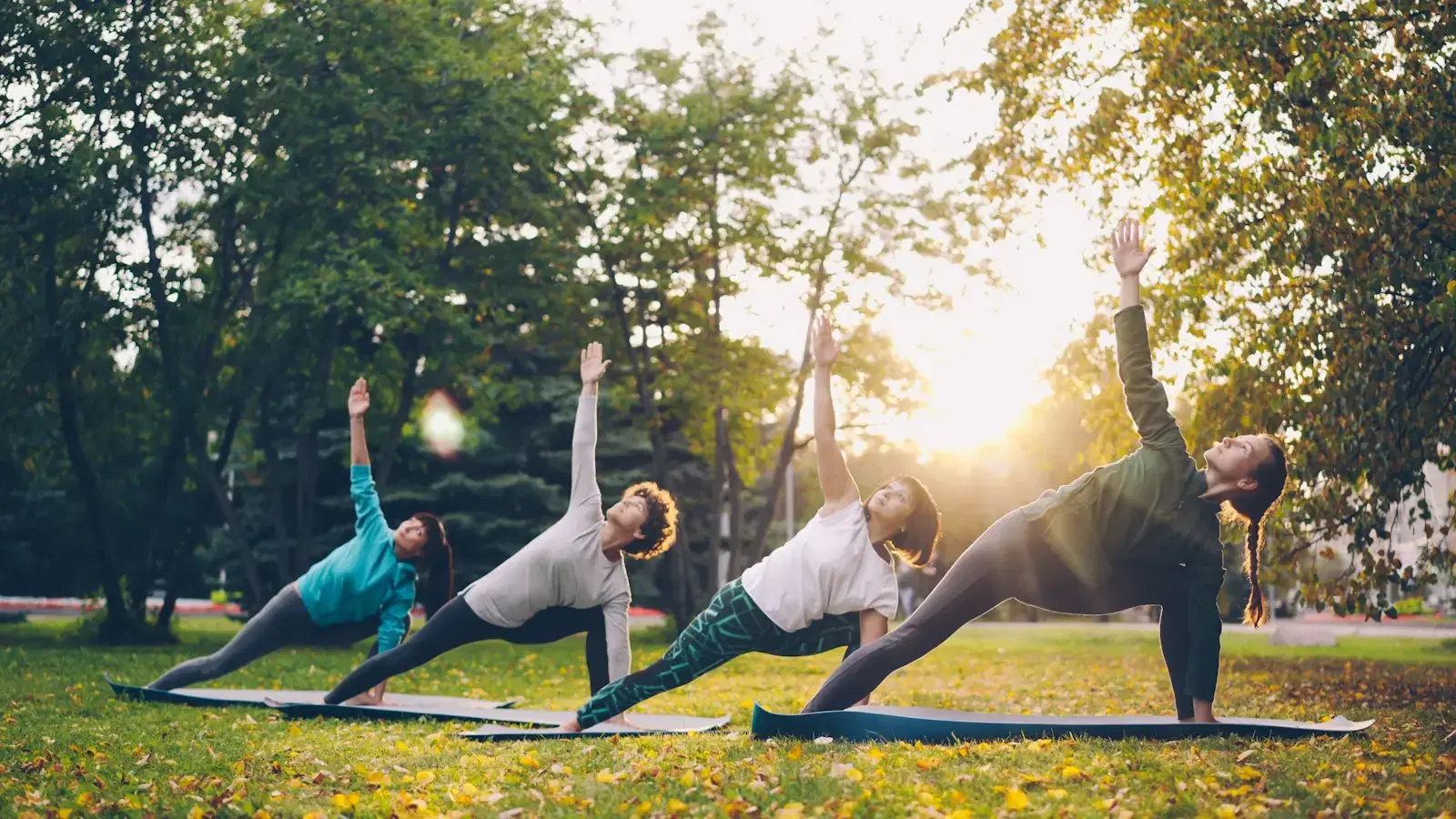 Four people holding a side-angle pose on mats in a park at sunset