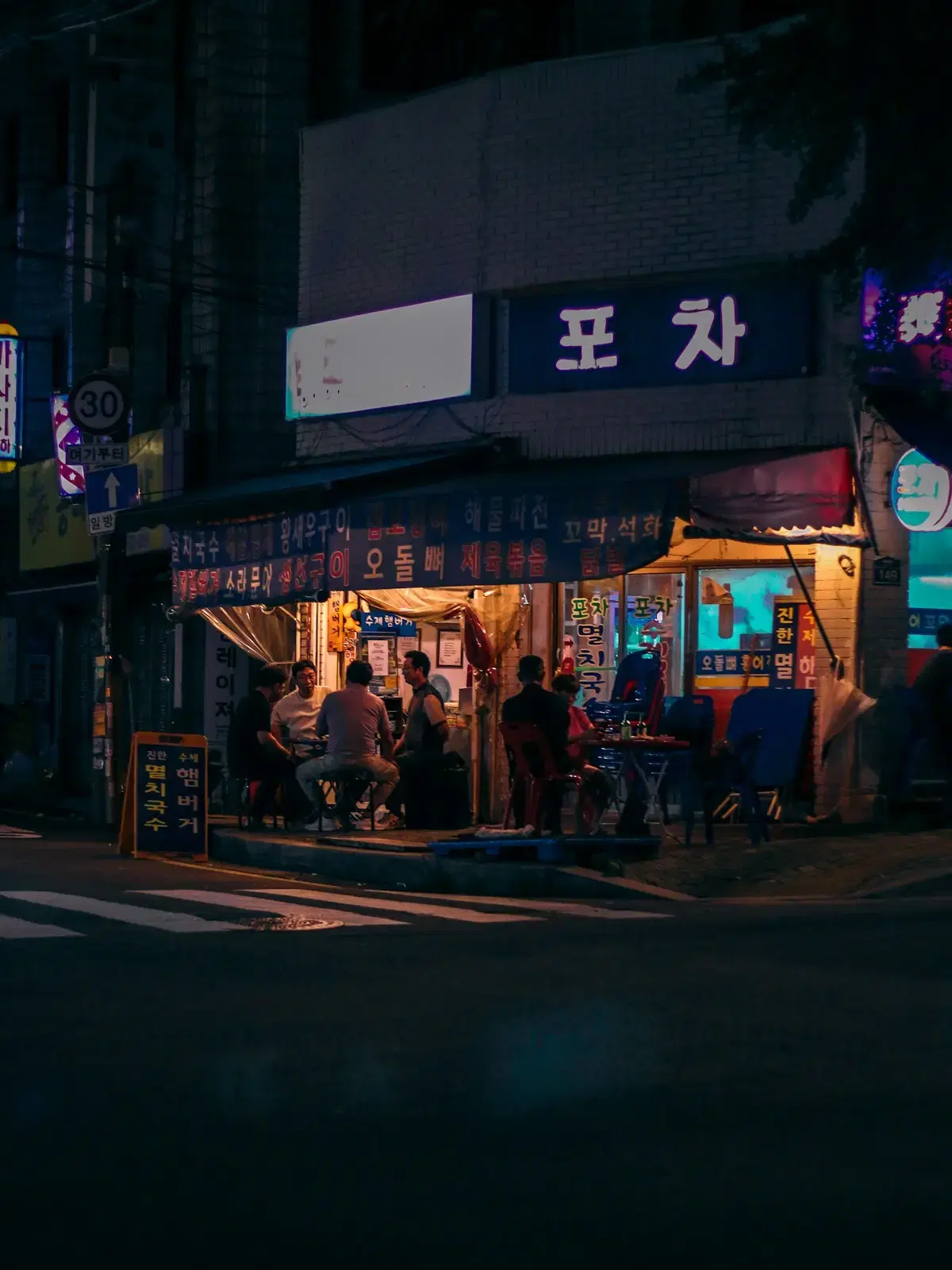 People at small tables outside a Korean street bar at night, its awning lit over an empty street
