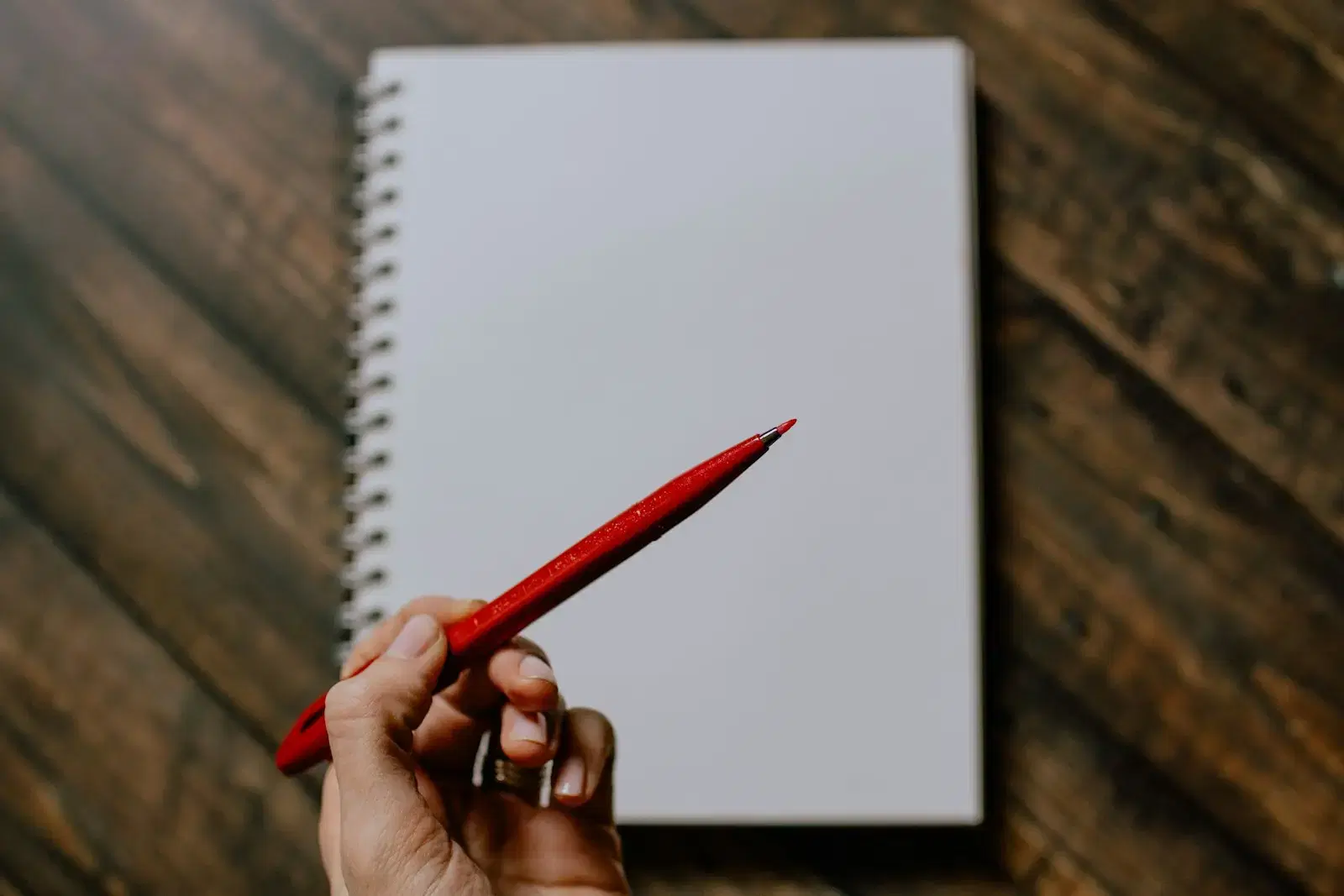 A hand holding a red pencil over a blank spiral notebook on a wooden table
