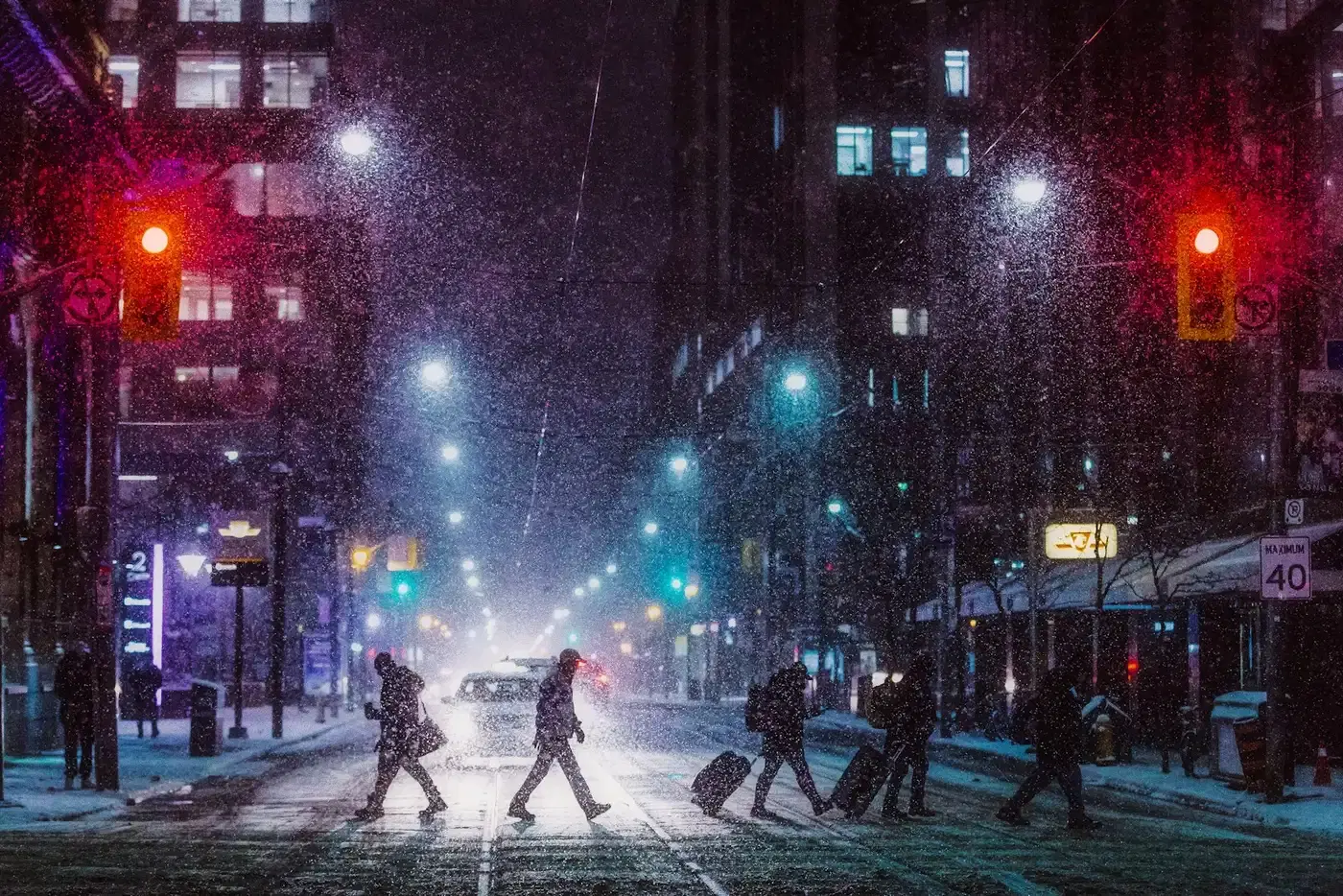 People crossing a city street at night in falling snow under red traffic lights