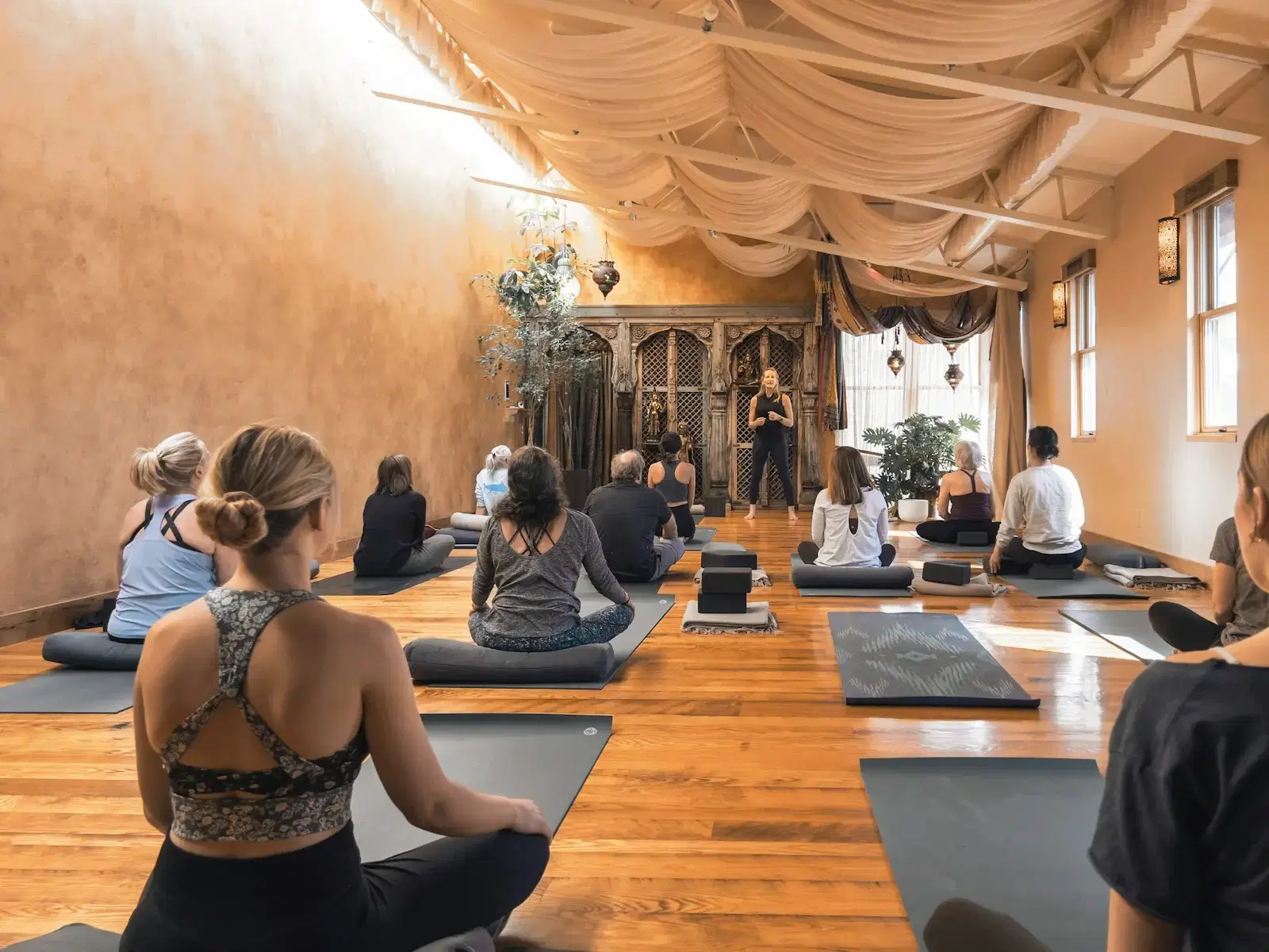 A yoga class seated on mats in a warm, wood-floored studio under draped fabric